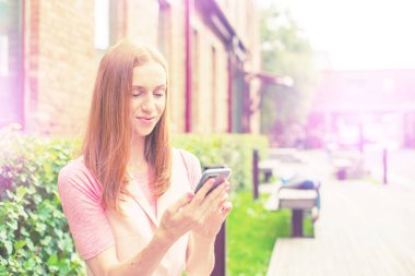 Güzel genç kız ahşap bir bankta oturuyor açık bir cep telefonunun üzerinde gülümsüyor. Güneşli bir gün yeşil çalılar yaşam tarzı seçici odaklanma tonu.