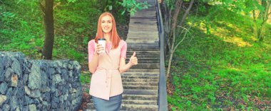 Afiş A güzel kızıl saçlı girl merdivenlerden aşağı gider ve bir kağıt bardak kahve içiyor. Park yaşam tarzı kahvaltı