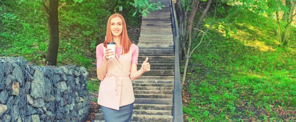 Afiş A güzel kızıl saçlı girl merdivenlerden aşağı gider ve bir kağıt bardak kahve içiyor. Park yaşam tarzı kahvaltı