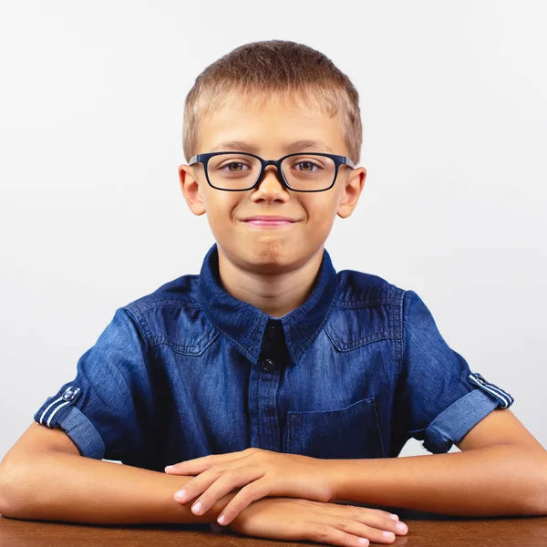 Uno scolaretto con una camicia blu seduto al tavolo. Ragazzo con gli occhiali su sfondo bianco Concetto torna a scuola — Foto Stock