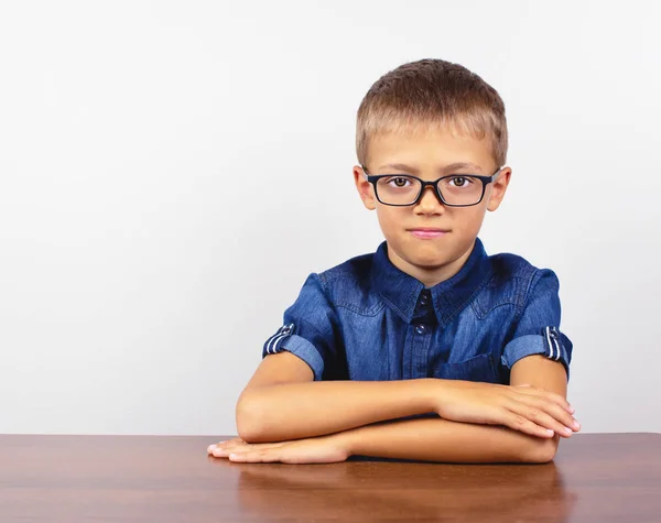 Uno scolaretto con una camicia blu seduto al tavolo. Ragazzo con gli occhiali su sfondo bianco Concetto torna a scuola — Foto Stock