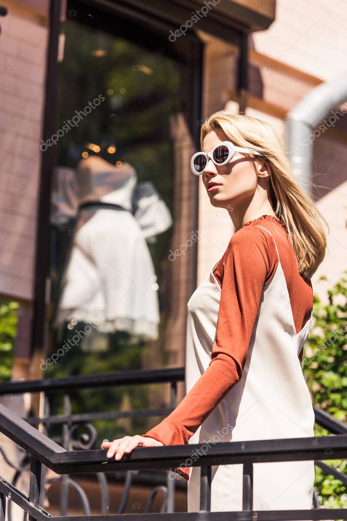 beautiful young woman in sunglasses looking away while standing near fashionable boutique