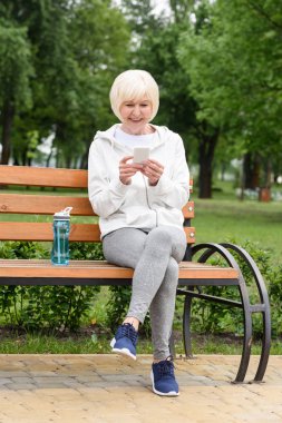 Yaşlı kadın ile spor şişe su bankta otururken smartphone kullanarak spor giyim