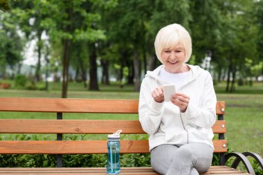 üst düzey kadın spor şişe su ile bankta otururken Smartphone kullanarak