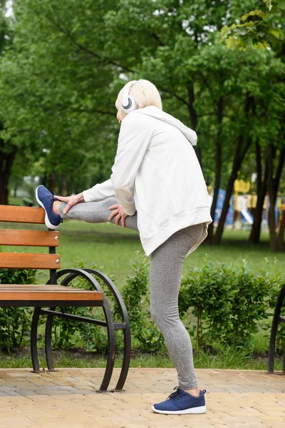 公園のベンチに近いストレッチ ヘッドフォンでシニアの運動女性  — 無料ストックフォト