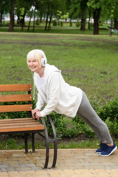 Seniorin Mit Kopfhörer Macht Liegestütze Nahe Bank Park — Stockfoto