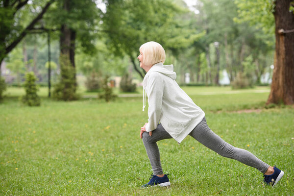 senior sportswoman exercising and doing lunges on lawn in park