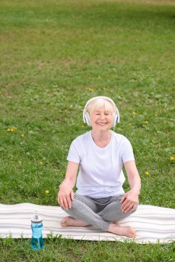 mutlu kadın kıdemli yoga mat parkta otururken kulaklık ile müzik dinleme 