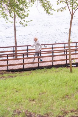 senior sportswoman jogging on wooden path in pak near lake clipart