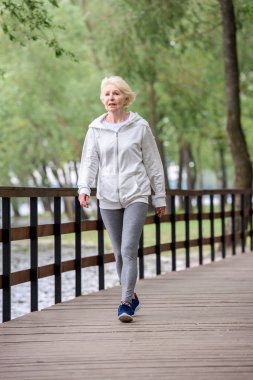 senior woman in sportswear walking on wooden path in park near river clipart