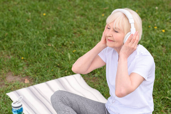 happy senior woman listening music with headphones while sitting on yoga mat on lawn 