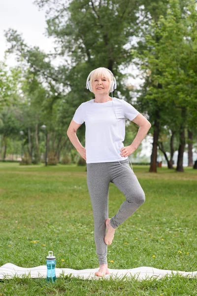 在公园瑜伽垫练习耳机的资深女瑜伽士 — 图库照片