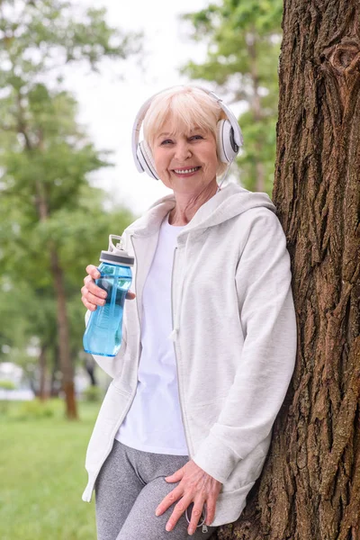 Smiling Senior Sportswoman Sport Bottle Listening Music Headphones Park — Stock Photo, Image