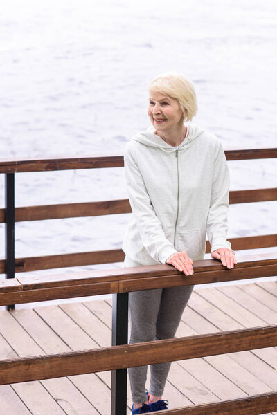 happy elderly woman standing near railings on wooden path near river 