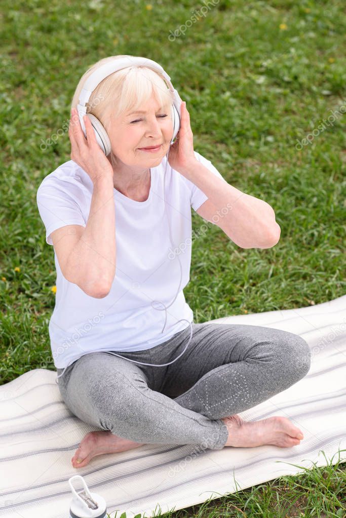 senior woman listening music with headphones while sitting on yoga mat