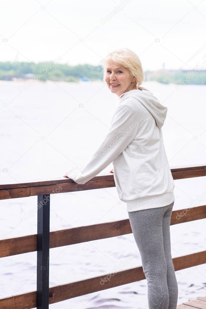 happy senior woman standing at wooden railings near river 