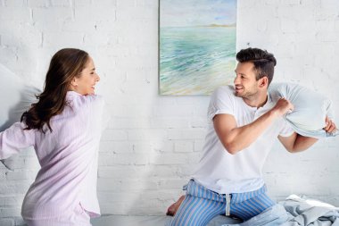 young happy girlfriend and boyfriend having pillow fight in bedroom  clipart