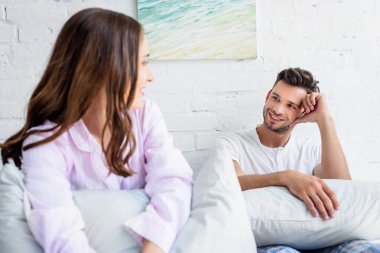 Genç çift pijama yatak yastık ile otururken birbirine bakarak gülen 