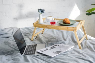 laptop with blank screen and breakfast with croissant and coffee on tray on bed in morning clipart