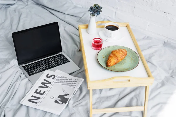 laptop, newspaper and breakfast with croissant and coffee on tray on bed in morning