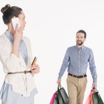 Woman with credit card talking on smartphone while husband carrying shopping bags isolated on white