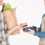 Vista parcial de la mujer con el paquete de papel con la comida que da tarjeta de crédito al asistente de la tienda con el lector de tarjetas aislado en blanco