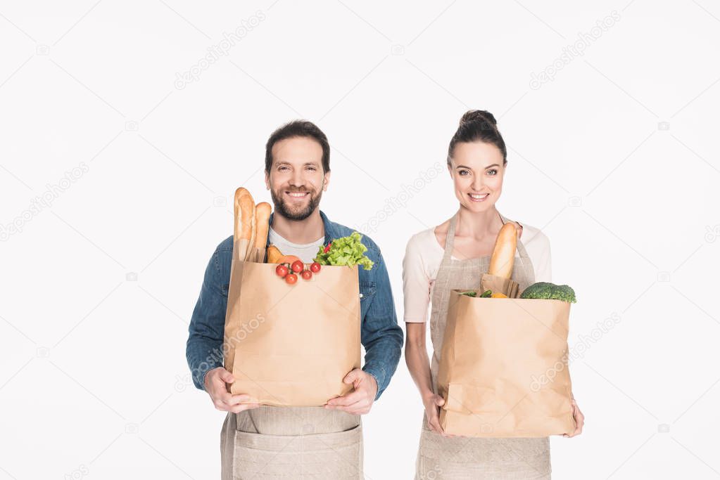 portrait of cheerful shop assistants in aprons holding paper packages with grocery isolated on white