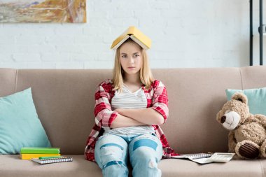 sad teen student girl with book on head sitting on couch at home and looking at camera clipart