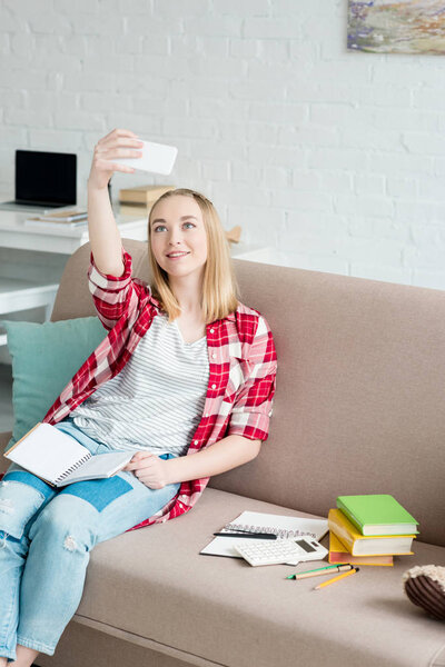 happy teen student girl with notebook sitting on couch and taking selfie