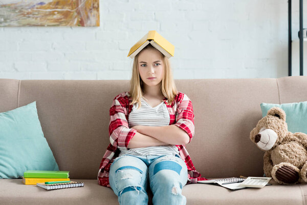 sad teen student girl with book on head sitting on couch at home and looking at camera