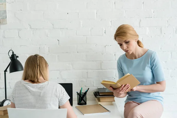 Moeder Leesboek Buurt Van Dochter Terwijl Werken Met Laptop — Stockfoto