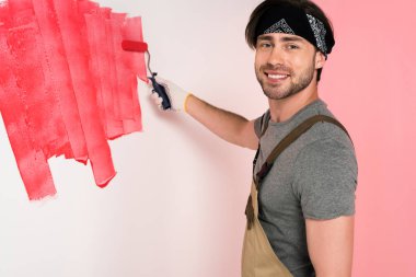 smiling man in working overall and headband painting wall in red by paint roller  clipart