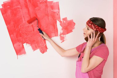kafa bandı ve genel smartphone ve boyama duvar kırmızı boya silindiri tarafından konuşurken çalışma genç kadın yan görünüm 
