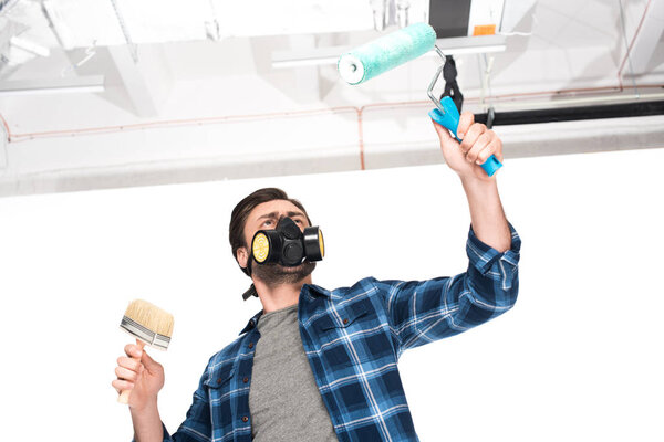 low angle view of man in respirator working by paint roller and paint brush 
