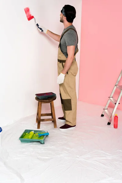 young man in working overall and headband painting wall in red by paint roller near chair with roller tray