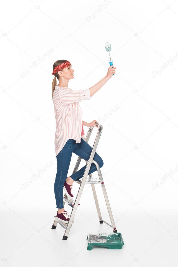 side view of woman in headband standing on ladder and painting by paint roller isolated on white background 