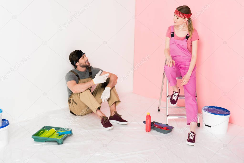 young couple in working overalls resting in room with ladder, paint tins, roller trays with paint rollers 