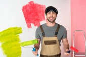 Junger lächelnder Mann im Arbeitsanzug mit Farbwalzen vor bemalter Wand 