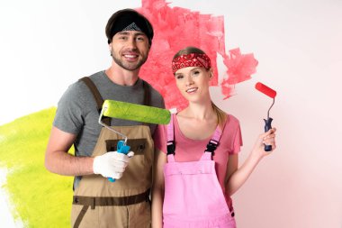 Genç çift iş yarı tulumlar ve saç bantları ile ayakta silindirler boyalı duvar önünde boya 