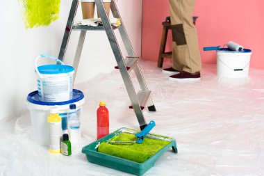 close up view of paint tins, bottles, ladder with coffee cups and protective gloves, roller tray with paint roller and man working behind  clipart