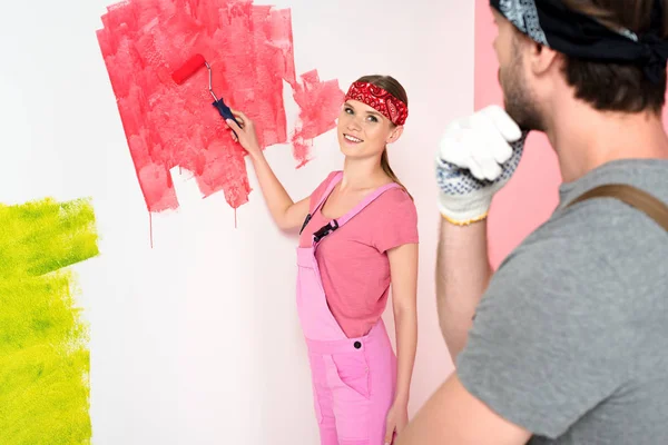 thoughtful man with hand on chin looking at girlfriend while she painting wall in red paint roller