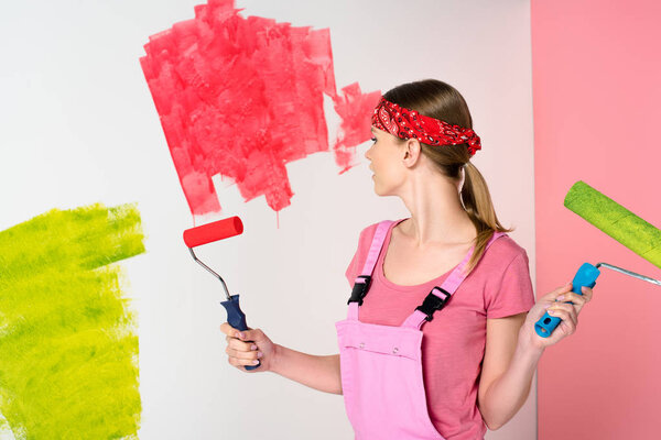 young woman in headband and working overall holding paint rollers in front of painted wall