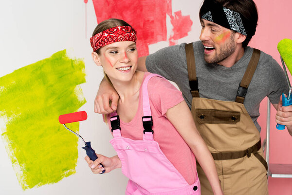 happy young couple in working overalls with painted faces holding paint rollers in front of painted wall 