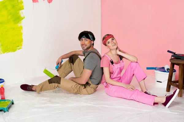 tired young couple in working overalls sitting on floor surrounded by painting tools