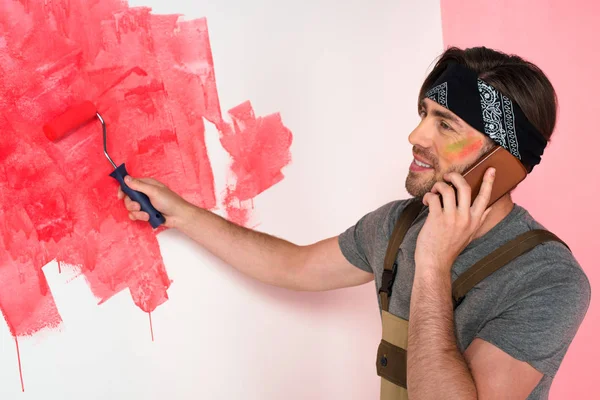 smiling man talking on smartphone and painting wall in red by paint roller