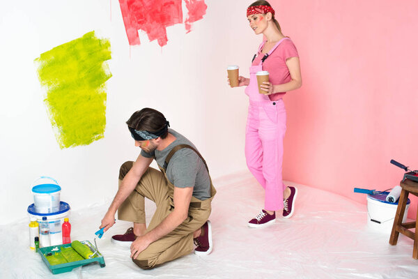 woman with paper cups of coffee approaching to boyfriend while he dipping paint roller into roller tray 