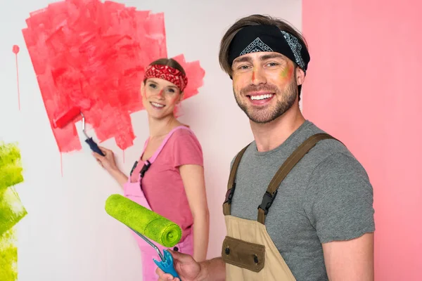smiling young couple in working overalls with painted faces holding paint rollers