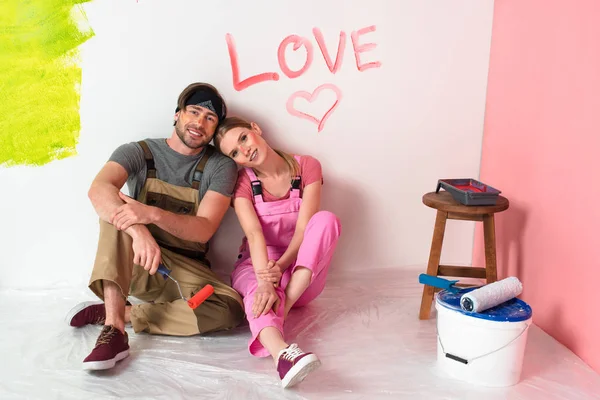 young couple in working overalls sitting near wall with lettering love and heart symbol