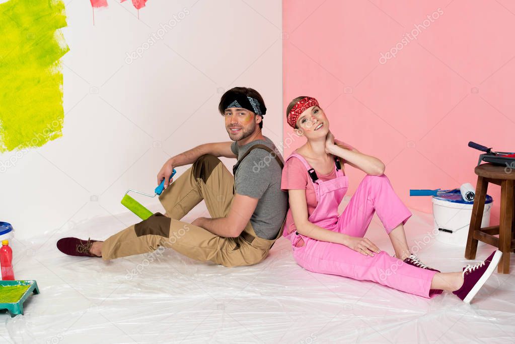 tired young couple in working overalls sitting on floor surrounded by painting tools 
