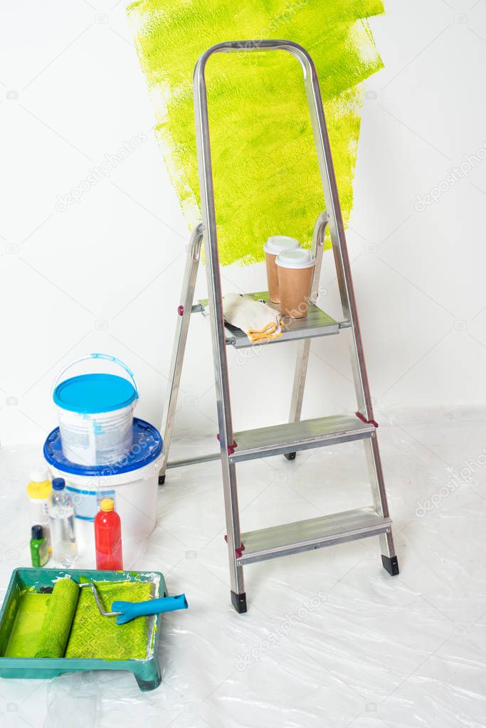 close up view of paint tins, bottles, roller tray with paint roller, ladder with protective gloves and paper cups of coffee 